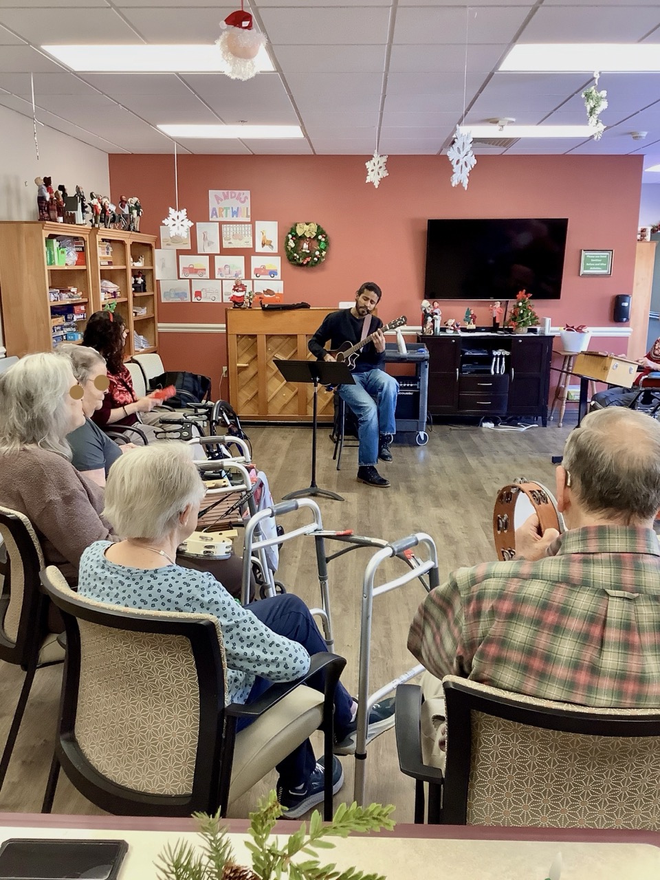 A music therapist playing guitar for a seated semi-circle of older adults during a holiday session, one shaking a tambourine