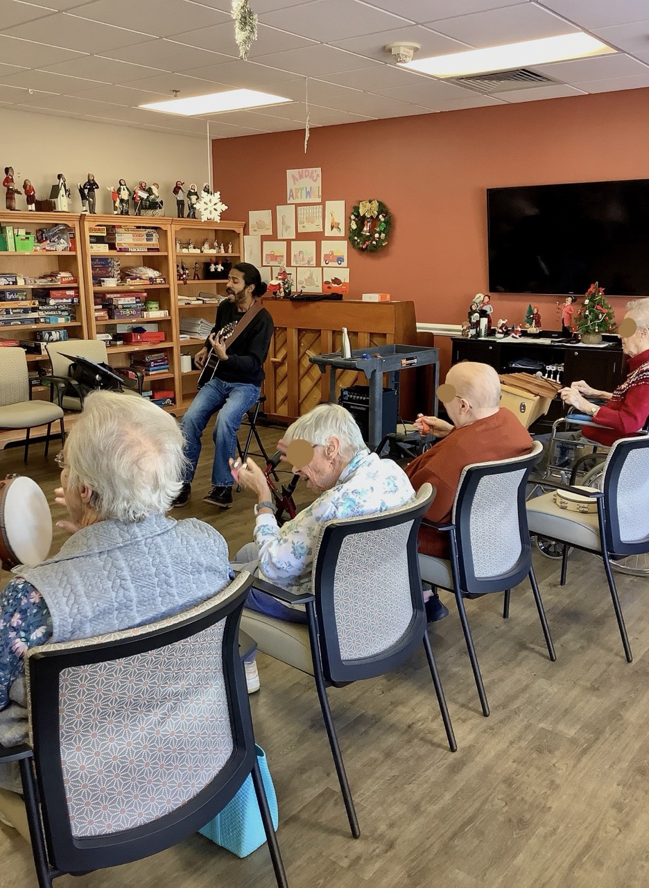A music therapist playing guitar for a group of older adults seated in a semicircle, some holding tambourines