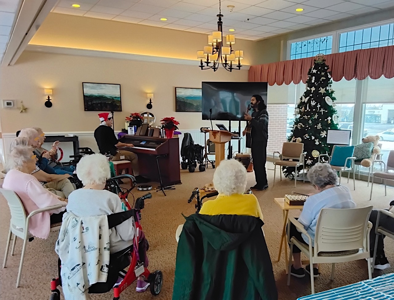 A music therapist singing and playing guitar beside a pianist for a group of older adults during a holiday session